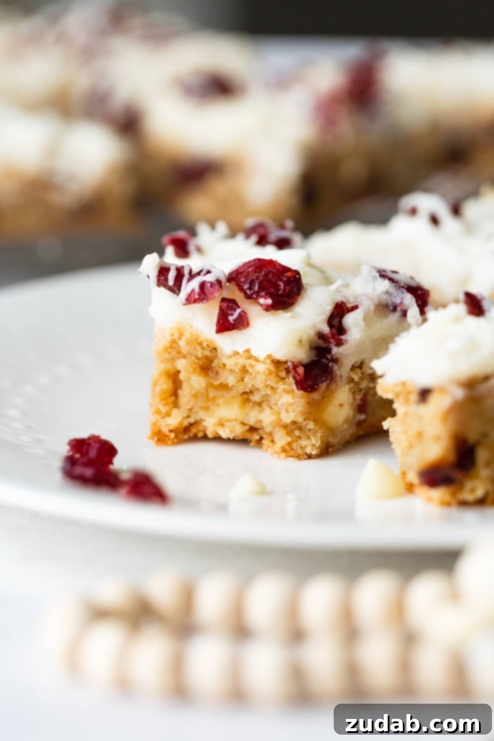 Beyaz Çikolatalı Kızılcık Keyfi 4 A close-up photo of a single, delectable cranberry bliss bar with a bite taken out of it, showcasing its soft, chewy interior filled with craisins and white chocolate chips, resting on a pristine white dessert plate.
