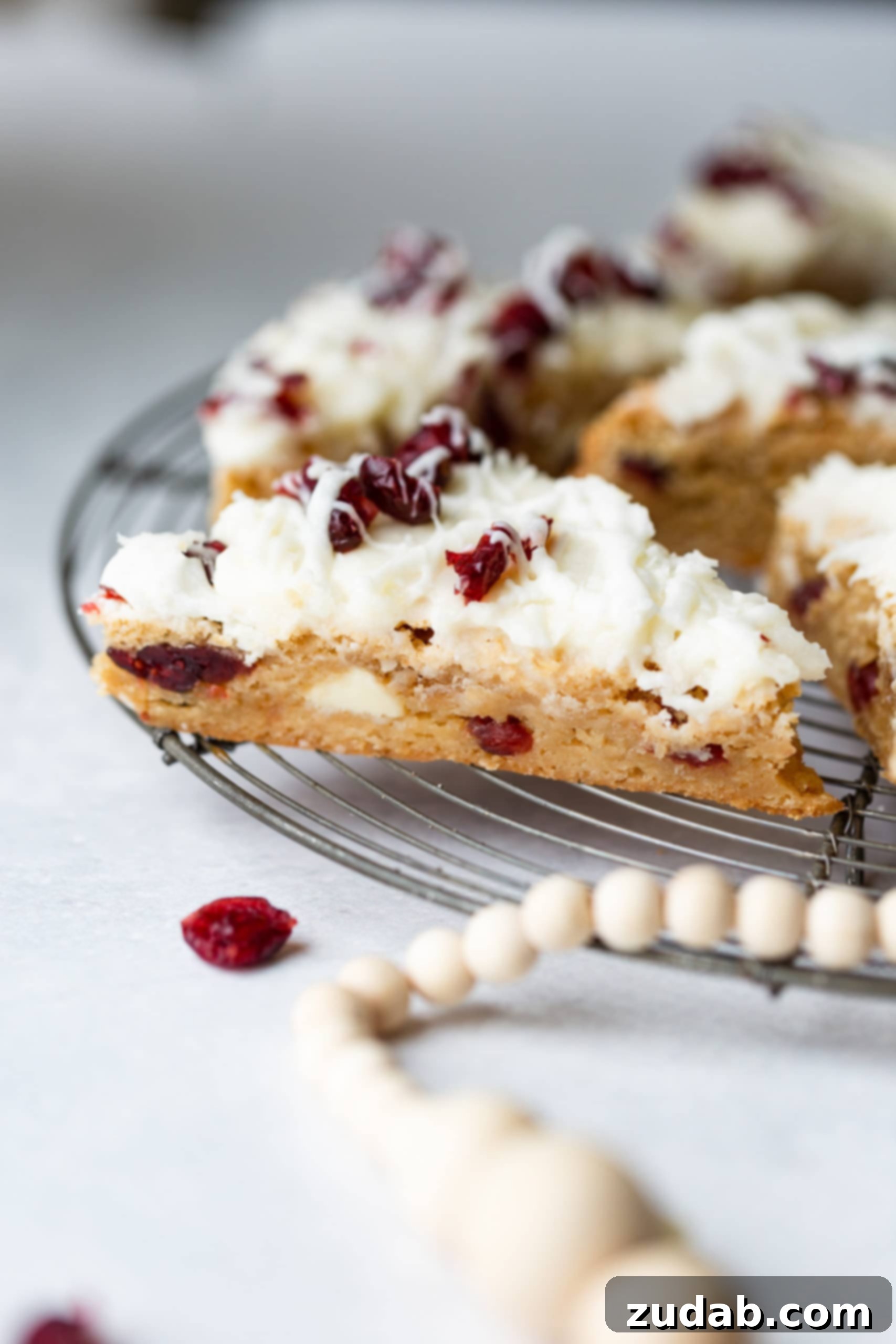 Beyaz Çikolatalı Kızılcık Keyfi 5 A circular cooling rack generously filled with triangular cranberry white chocolate bars, richly studded with craisins and white chocolate chips, and elegantly topped with a thick layer of cream cheese frosting.