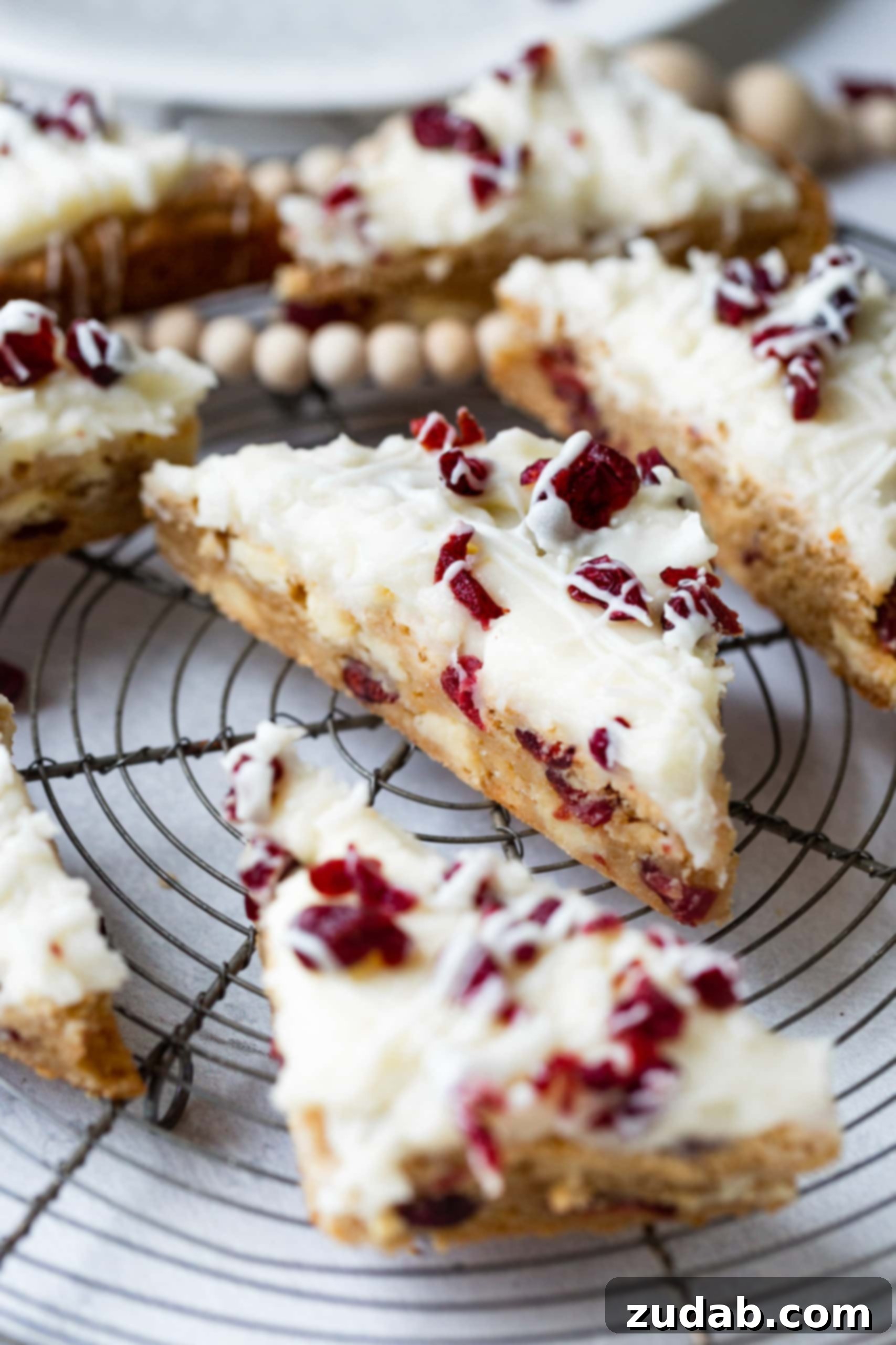 Beyaz Çikolatalı Kızılcık Keyfi 6 A collection of perfectly cut, triangular white chocolate cranberry bliss bars elegantly arranged on a circular cooling rack, creating a festive and inviting dessert display.