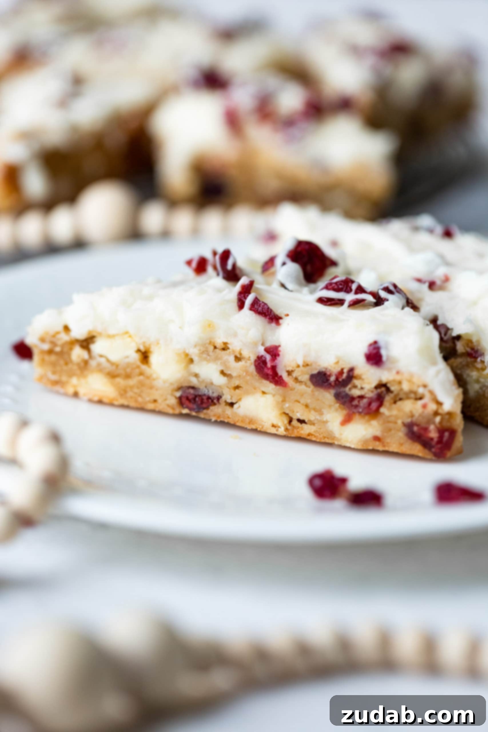 Beyaz Çikolatalı Kızılcık Keyfi 7 A single, perfectly frosted white chocolate cranberry bliss bar, adorned with craisins and a delicate white chocolate drizzle, presented elegantly on a white dessert plate.