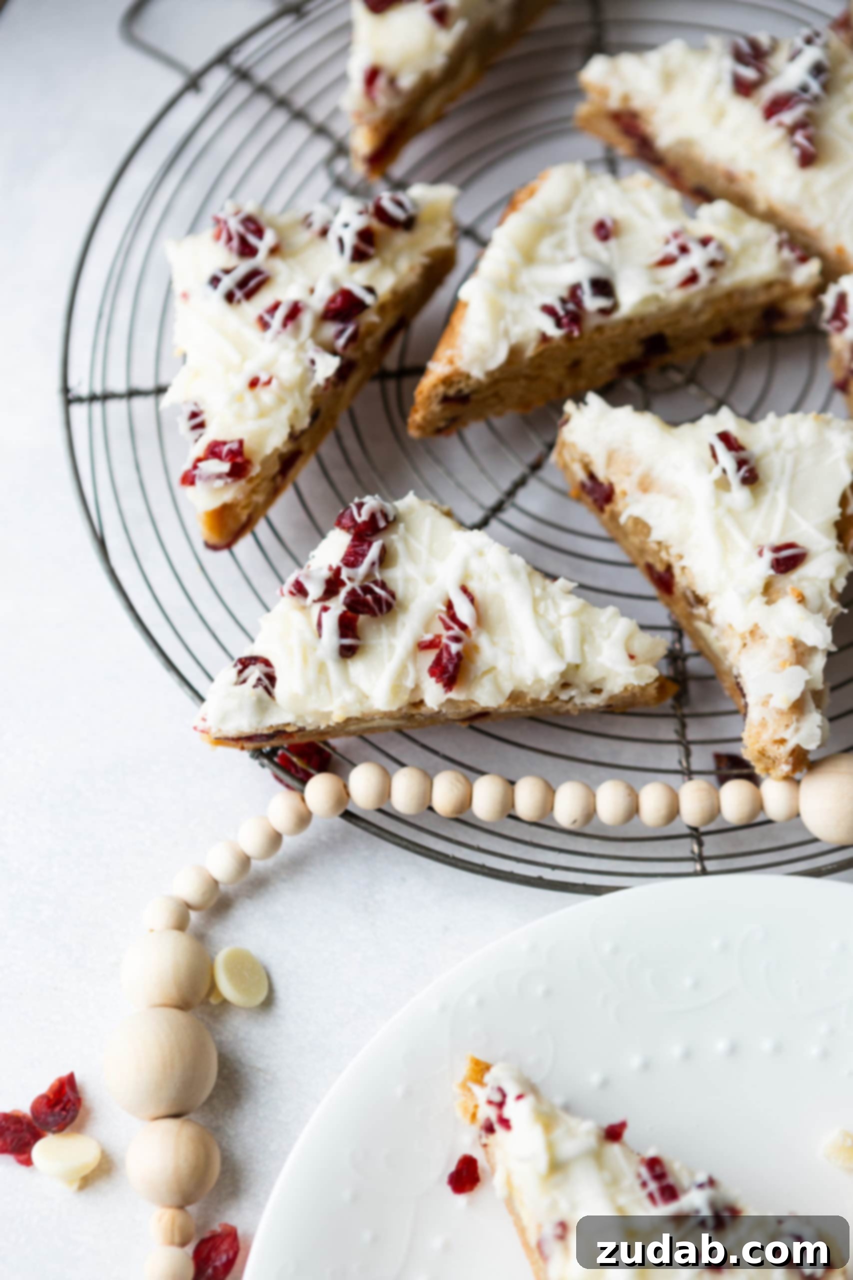 Beyaz Çikolatalı Kızılcık Keyfi 8 Several triangle-cut white chocolate cranberry bliss bars arranged on a circular cooling rack, accompanied by a string of decorative beige beads, highlighting their festive appeal.