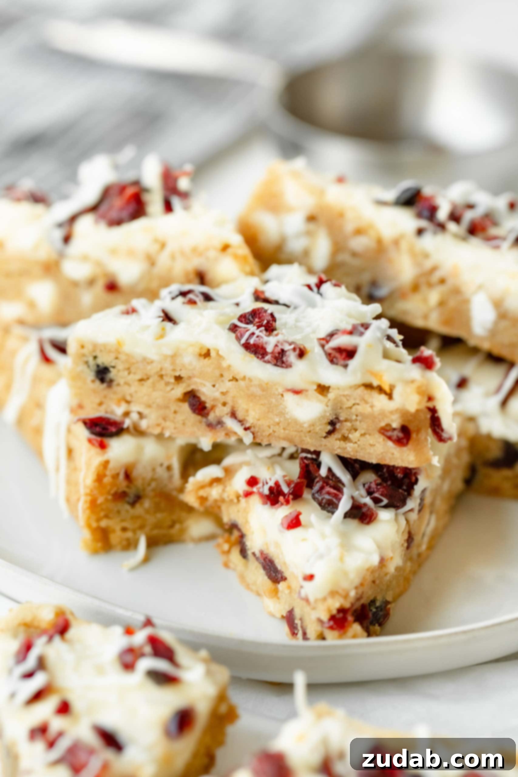 a photo of several orange cranberry bliss bars stack on each other. they are drizzled with a white chocolate drizzle and topped with dried cranberries.