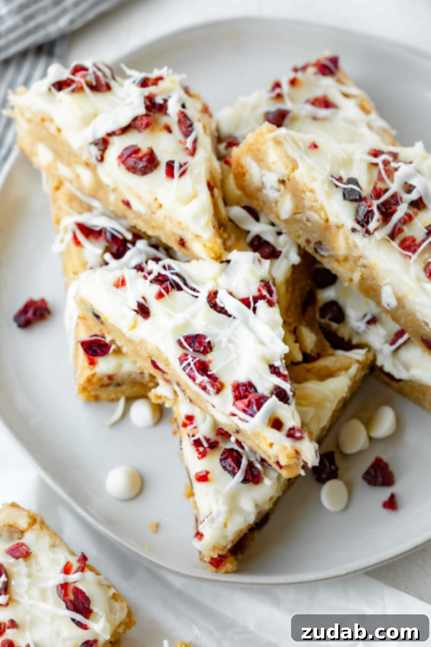 a photo of several orange cranberry bliss bars that have been cut into triangles and are stacked on each other. they are drizzled with a white chocolate drizzle and topped with dried cranberries.