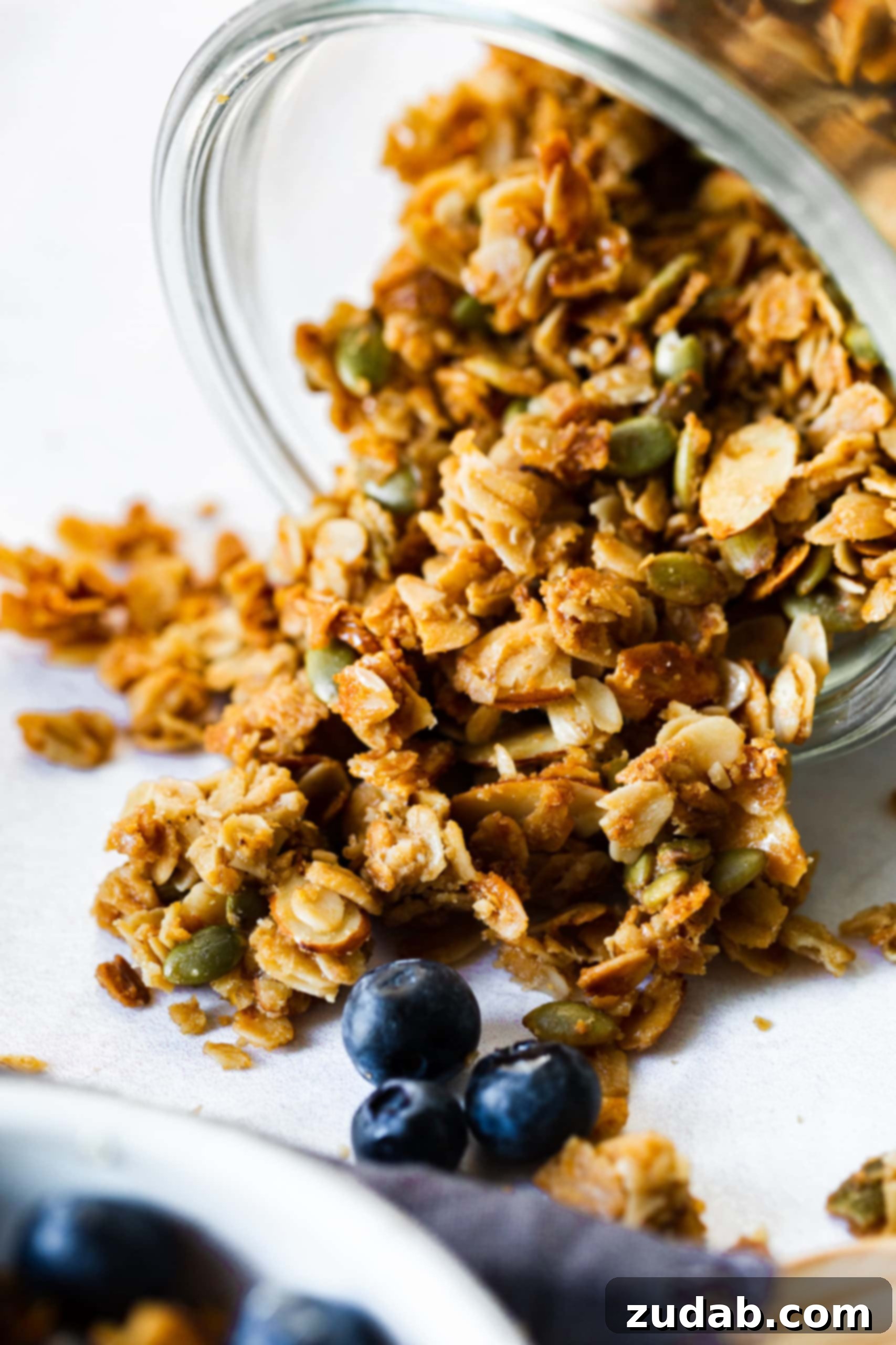 Your New Go-To Granola 2 a photo of homemade granola being poured out of a container with fresh blueberries in the foreground.