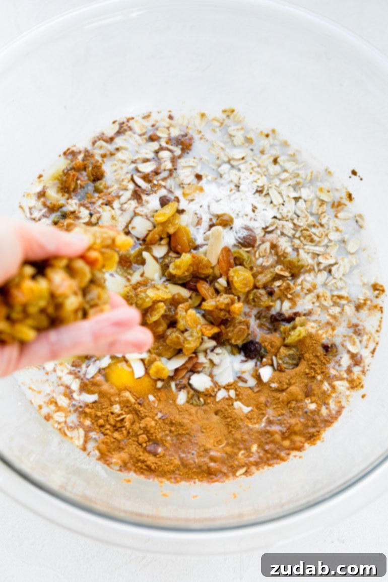 Wholesome Apple Fritter Baked Oats 4 A photo of a large glass bowl, rich with uncooked oatmeal, dried apple chunks, and aromatic spices, as a hand carefully adds golden raisins, ensuring an even distribution of flavors.