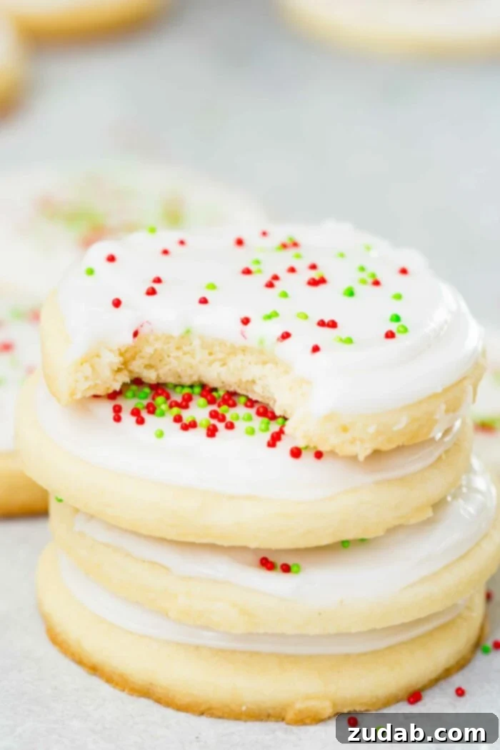 Festive Cookie Collection 8 A tempting stack of four sugar cookies, each adorned with creamy white frosting and festive red and green sprinkles. A bite has been taken out of the top cookie, revealing its soft interior.