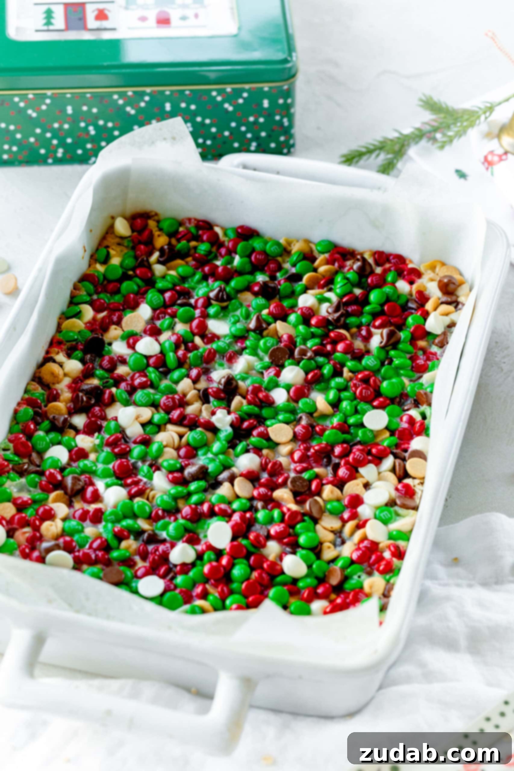 Christmas Magic Cookie Bars A photo of a rectangular white baking dish filled with baked Christmas magic bars, generously topped with Christmas-colored M&M's, white chocolate chips, peanut butter chocolate chips, and semi-sweet chocolate chips.