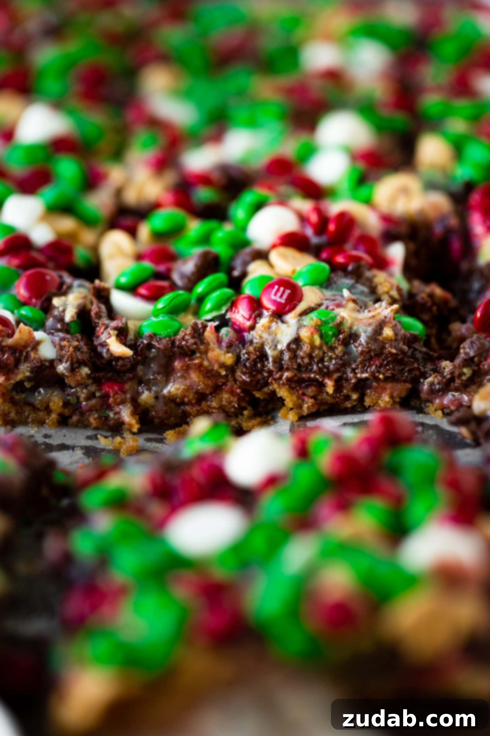 Christmas Magic Cookie Bars A photo of a magic 7 layer bar taken from the side, showcasing the distinct layers: a bottom graham cracker crust, a layer of oats, then sweetened condensed milk, and finally a generous topping of baking chips and holiday M&M's.
