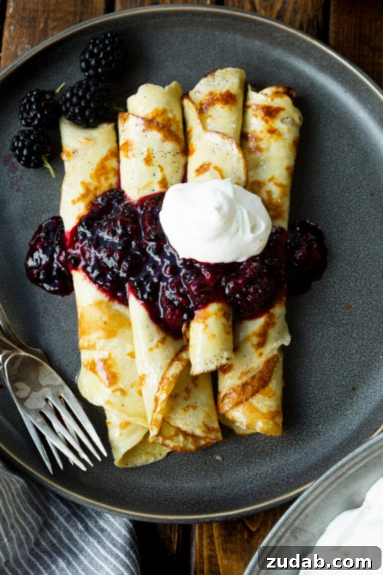 A beautifully arranged plate featuring several rolled Finnish pancakes, generously topped with a rich blackberry syrup and a dollop of fresh whipped cream, ready to be enjoyed.