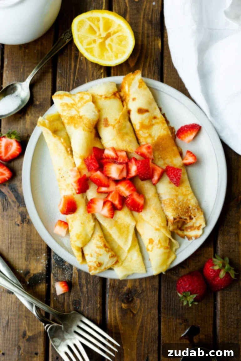 Classic Swedish Pancakes 3 A close-up photograph of four elegantly rolled Swedish pancakes, artfully placed on a white ceramic plate and generously sprinkled with bright red diced strawberries, emphasizing their golden-brown hue and tender appearance.