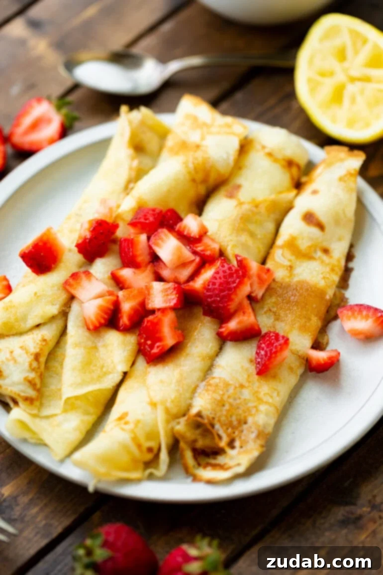 Classic Swedish Pancakes 5 A beautiful arrangement of four rolled Swedish pancakes resting on a white plate, artfully topped with a generous scattering of vibrant diced strawberries, presenting a fresh and appealing breakfast dish.