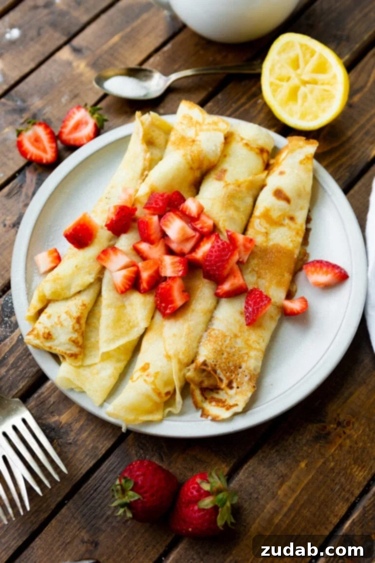 Classic Swedish Pancakes 6 A white ceramic plate holds four neatly rolled, golden Swedish pancakes, beautifully topped with a colorful scattering of diced strawberries, and a gleaming silver fork rests invitingly beside them.