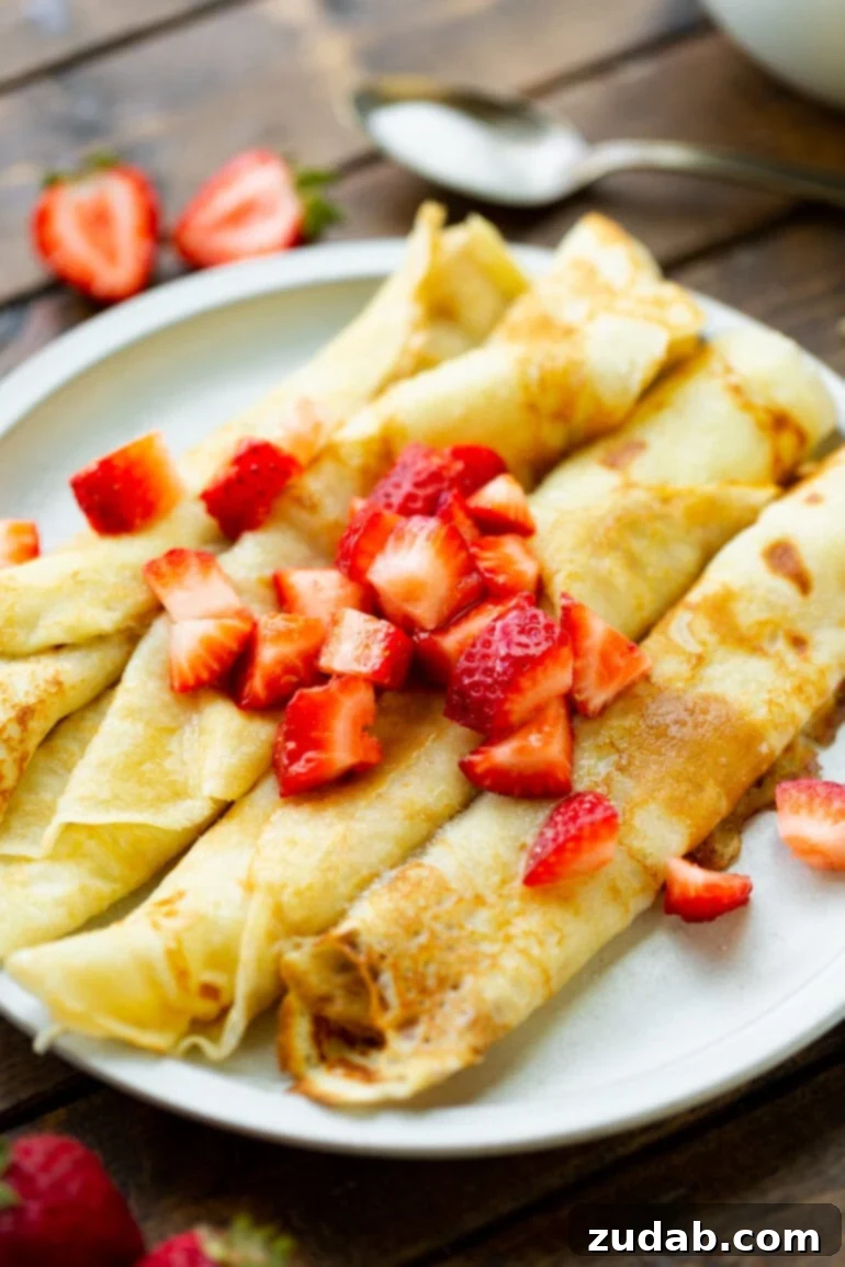Classic Swedish Pancakes 7 A delightful presentation of several golden-brown Swedish pancakes, skillfully rolled and neatly arranged on a white plate, generously adorned with freshly diced strawberries. This image captures the inviting and mouth-watering essence of a perfect breakfast.