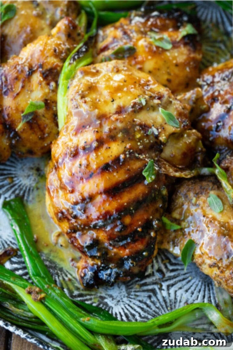 Maple Mustard Glazed Grilled Chicken Thighs 2 A close-up photo of a grilled chicken thigh covered in a vibrant maple mustard marinade, resting next to several other perfectly charred chicken thighs.