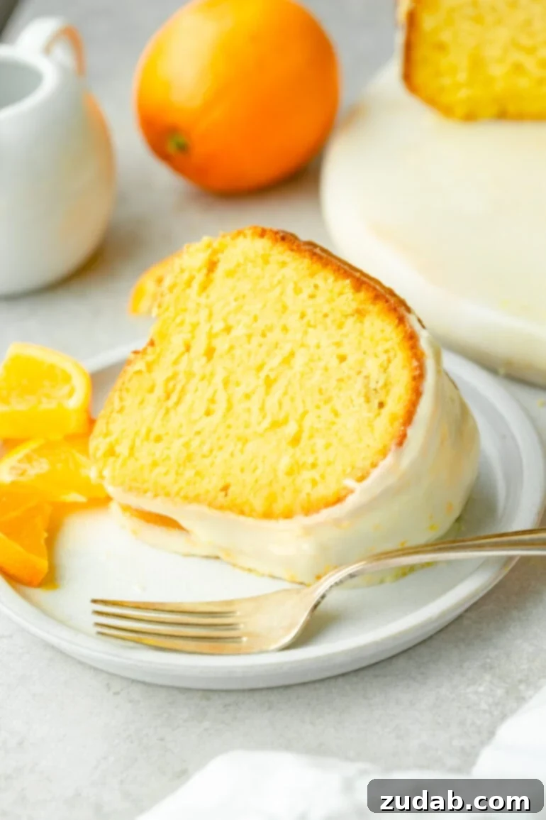 A slice of orange cake, drizzled with a smooth white glaze, rests on a white plate. A fork sits nearby, complemented by several orange slices.