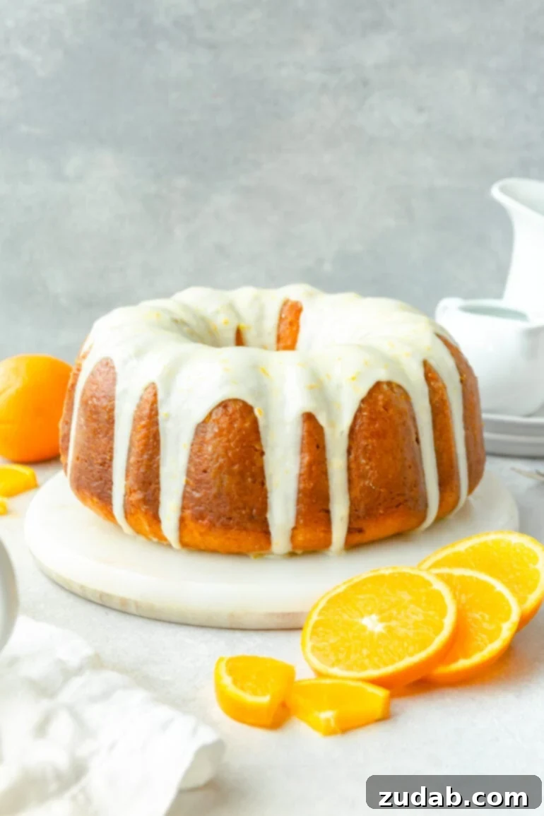a photo of a whole classic glazed orange bundt cake sitting on a white serving tray. Fresh orange slices surround the cake.