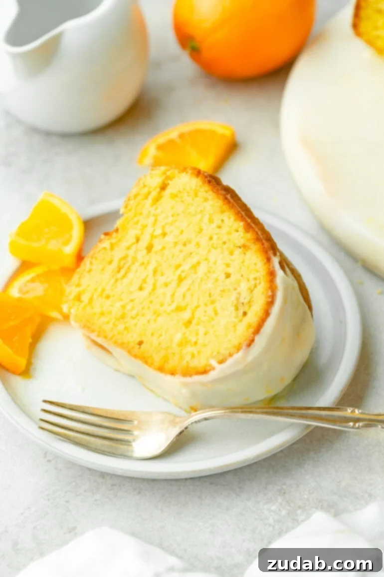 a photo of a slice of classic glazed orange bundt cake sitting on a white dessert plate with a silver spoon sitting next to it and small orange slices scattered around it.