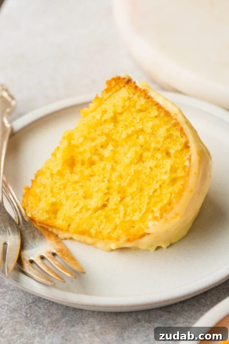 a photo of a piece of classic glazed orange bundt cake sitting on a small white plate with a silver fork sitting next to it.