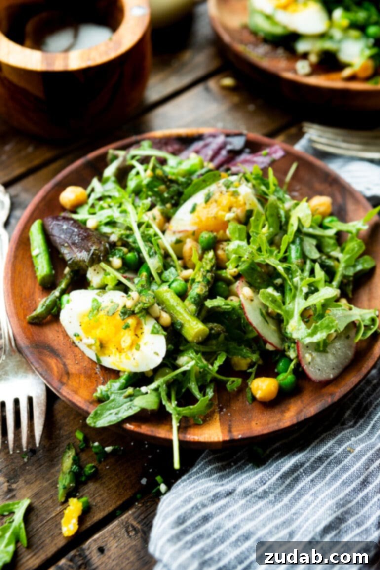 Effortless Springtime Greens 2 a photo of a wooden dinner plate full of fresh spring salad including greens, asparagus, hard boiled eggs, roasted chickpeas and topped with a lemon vinaigrette.