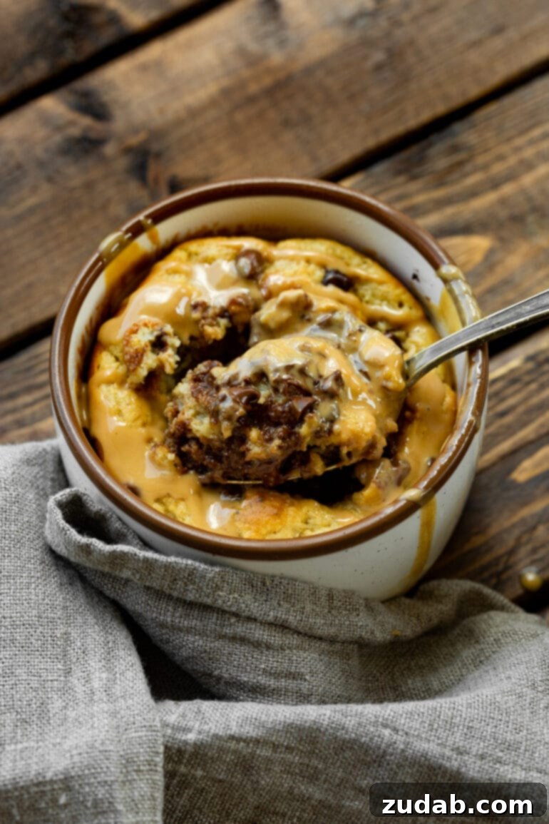 An appetizing close-up of a spoonful being skillfully scooped from the heart of a baked peanut butter chocolate chip oatmeal mug cake. The cake is beautifully topped with mini chocolate chips and a rich drizzle of peanut butter, highlighting its irresistible texture.