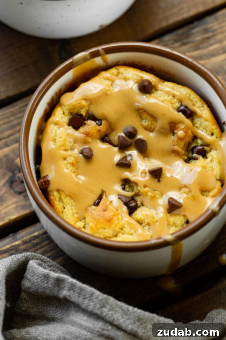 A top-down view of a perfectly baked oatmeal mug cake, generously topped with an array of mini chocolate chips and a luxurious drizzle of warm peanut butter, creating an inviting visual of this sweet and savory breakfast.