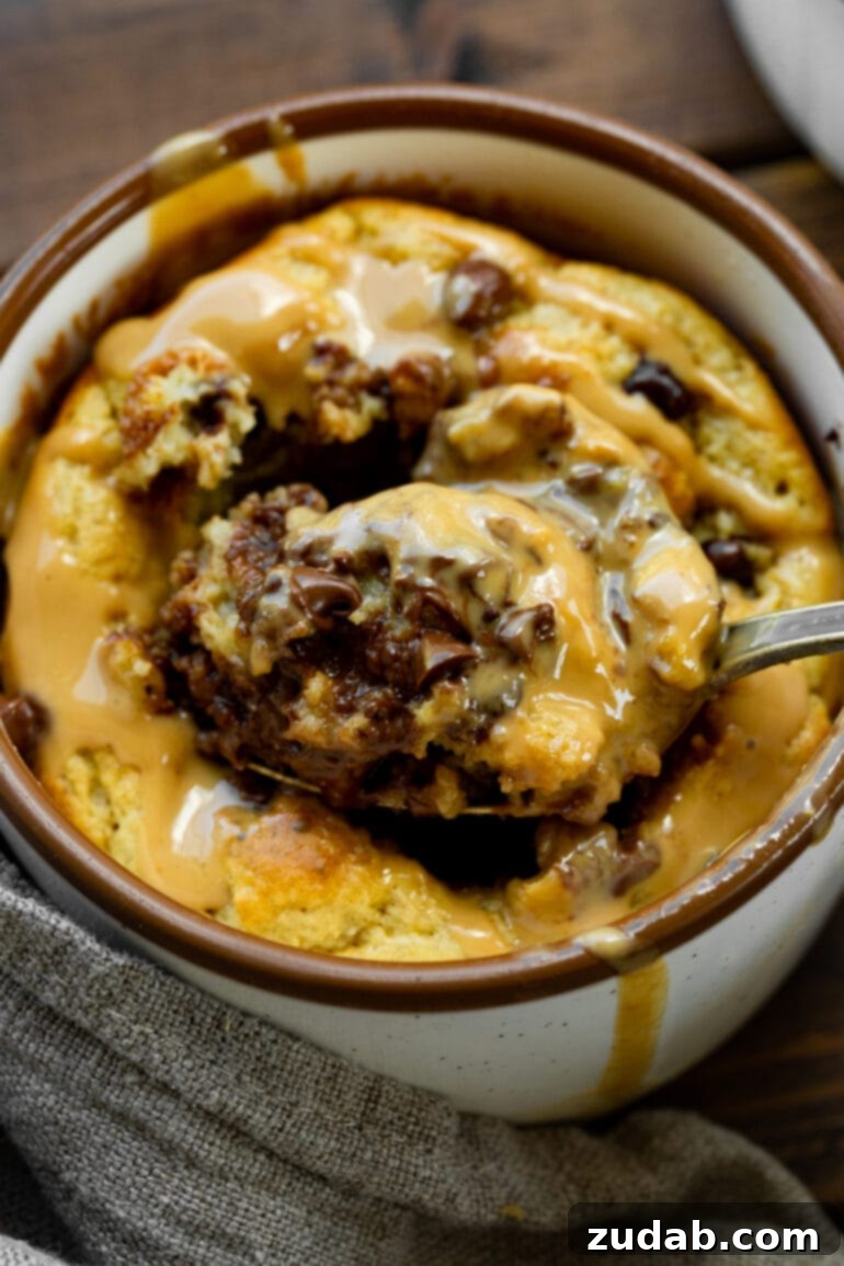 A tantalizing shot of a spoon expertly scooping out the soft, warm center of a baked oatmeal mug cake, lavishly topped with mini chocolate chips and a glistening drizzle of hot peanut butter, inviting a delicious taste.