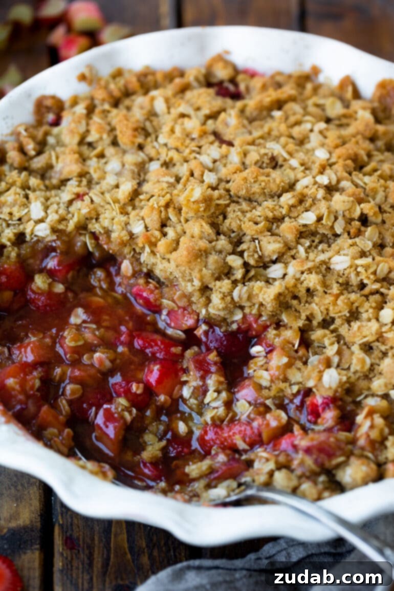 Golden Strawberry Rhubarb Crisp 5 A photo of a baked strawberry rhubarb crisp in a circular white ceramic baking dish, with a serving removed to reveal the juicy, bright red filling and golden, buttery oat topping.