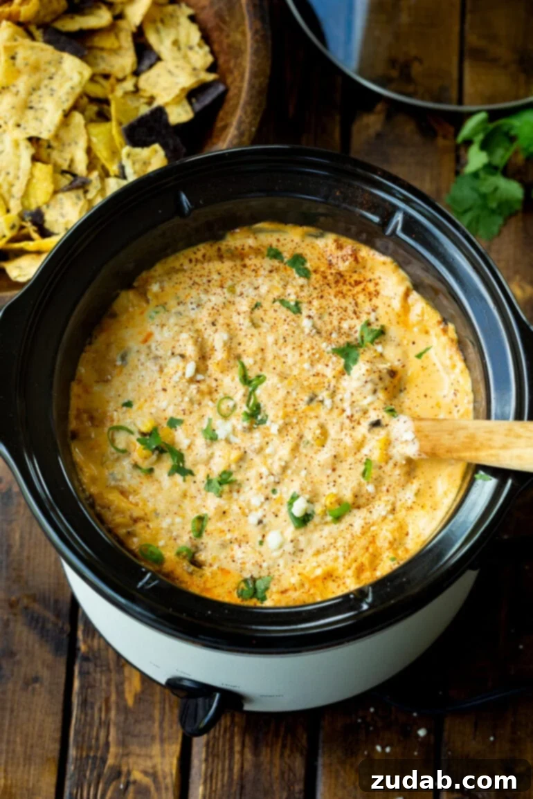 Creamy Slow Cooker Elote Dip 3 a photo of a small crockpot full of mexican street corn dip with a wooden spoon stuck into it.