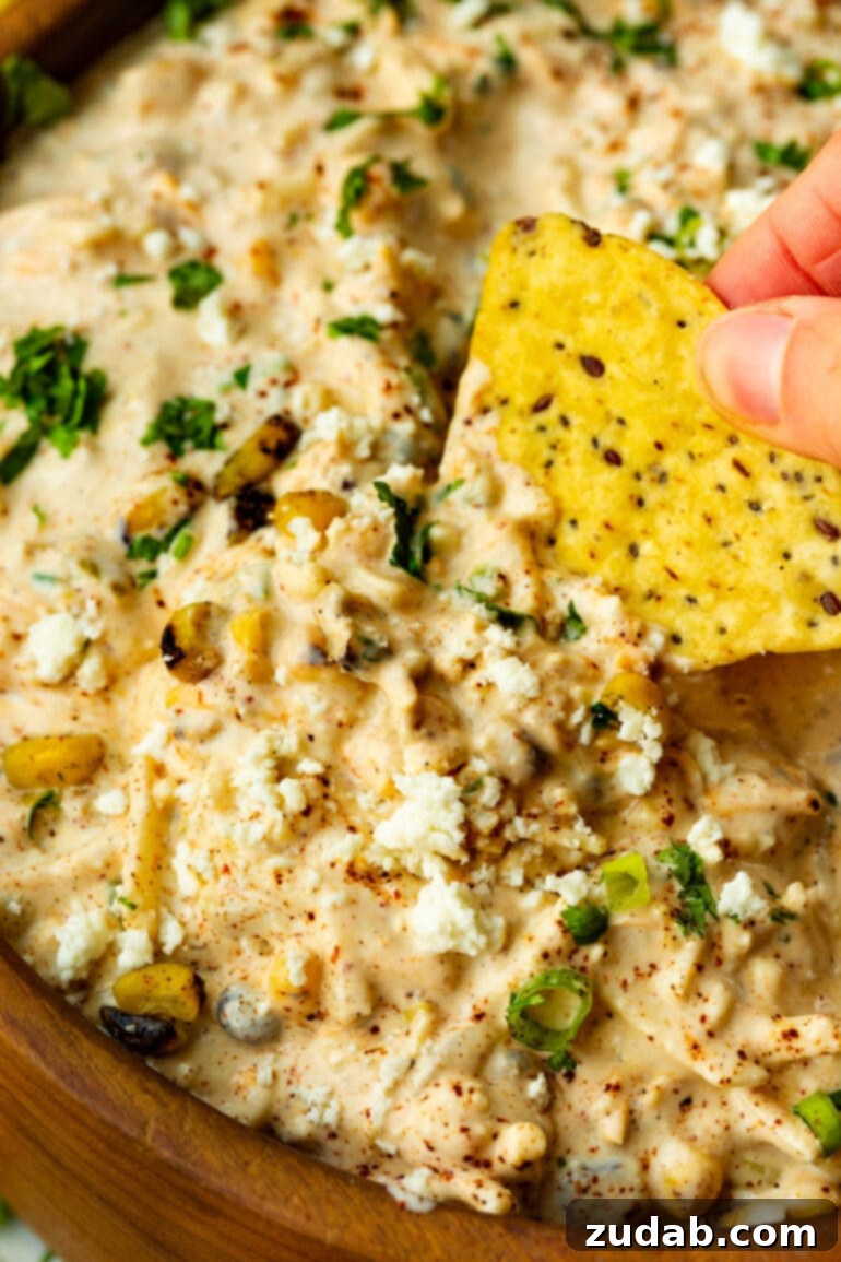 Irresistible Elote Dip 3 A close-up shot of a hand dipping a crispy tortilla chip into a generously filled bowl of vibrant Mexican corn dip, highlighting the creamy texture and colorful ingredients.