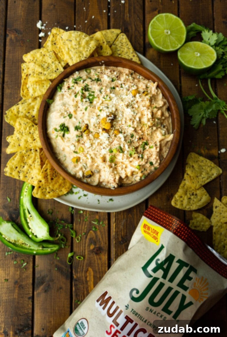 Irresistible Elote Dip 4 An overhead view of a large wooden bowl brimming with vibrant Mexican corn dip, surrounded by a generous array of golden tortilla chips and a bag of chips resting nearby. A halved jalapeno adds a touch of color.