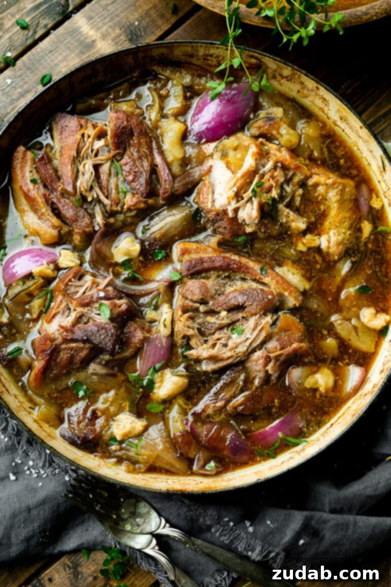 Sweet & Savory Apple Cider Braised Pork 2 a photo of chunks of braised pork butt in a large dutch oven sitting in a rich sauce full of apple slices, onion slices and fresh herbs.