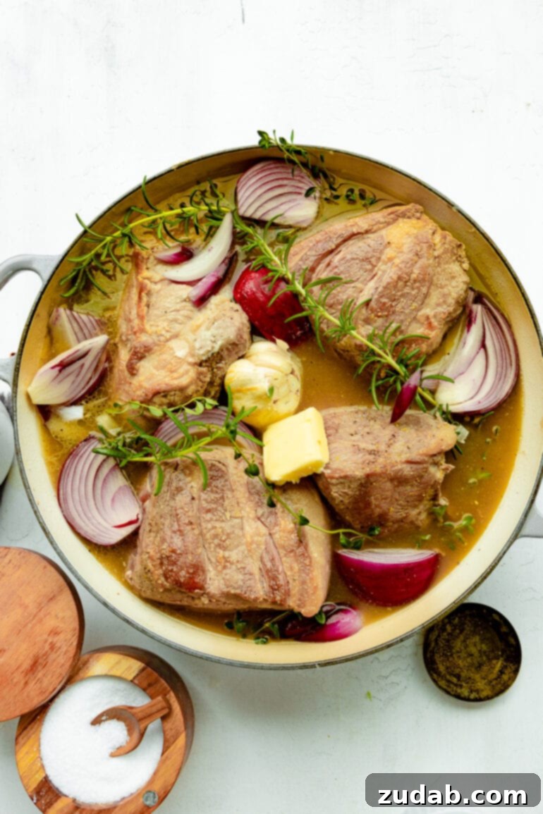 Sweet & Savory Apple Cider Braised Pork 3 a photo of a large dutch oven full of chunks of partially cooked pork butter, slices of red onion, fresh sprigs of rosemary and time all sitting in a braising liquid.