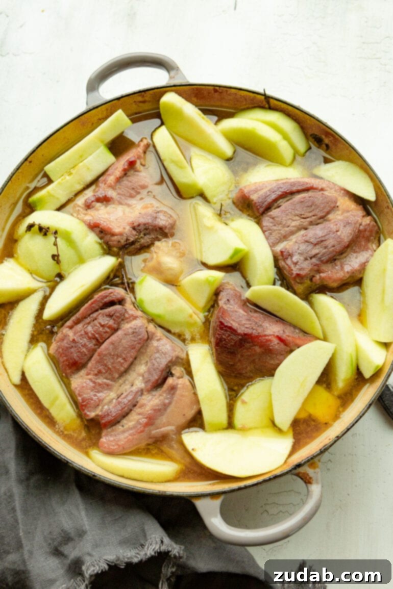Sweet & Savory Apple Cider Braised Pork 4 a photo of a large dutch oven full of partially cooked chunks of pork shoulder and apple slices all sitting in a braising liquid.