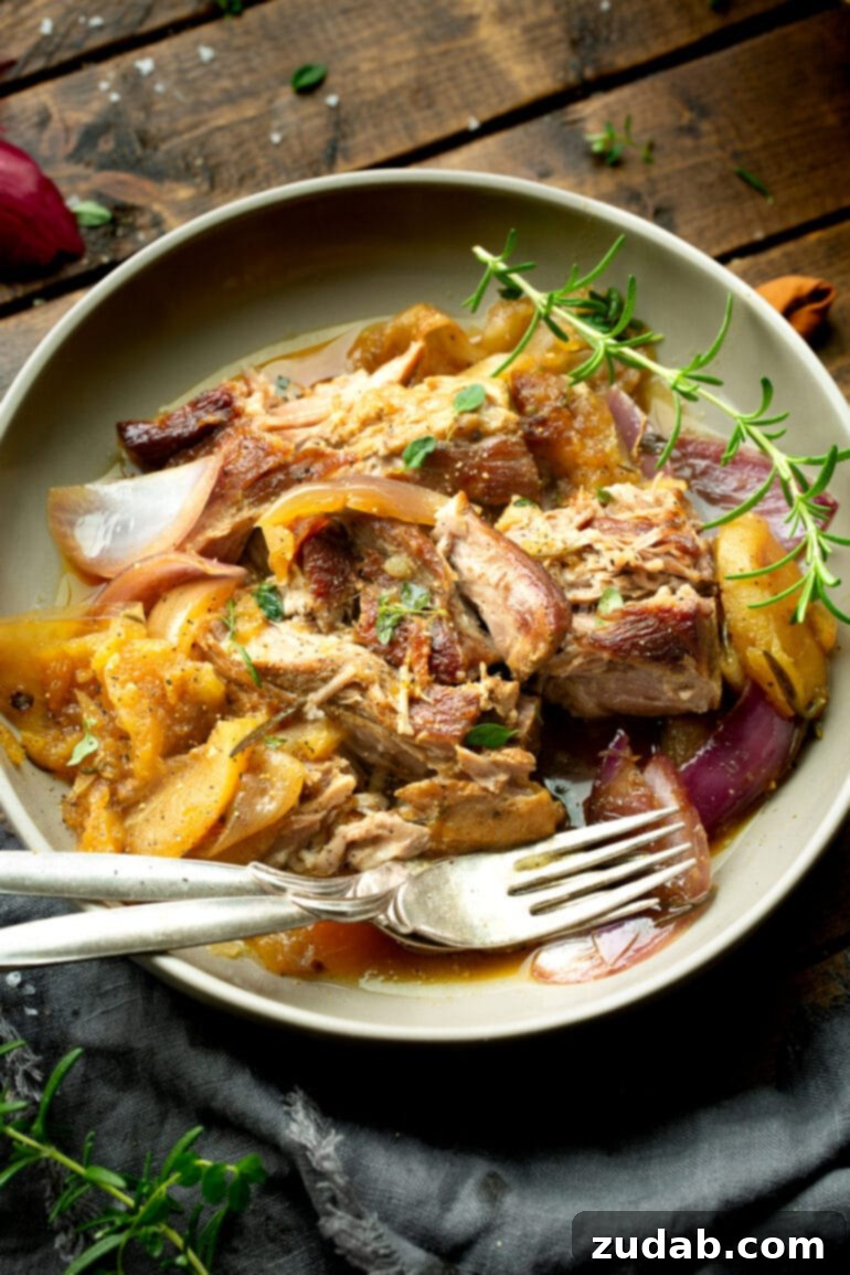Sweet & Savory Apple Cider Braised Pork 6 a photo of a large serving dish full of pieces of braised pork, apple slices, red onion slices and topped with fresh herbs with two forks sitting on the side of the plate.