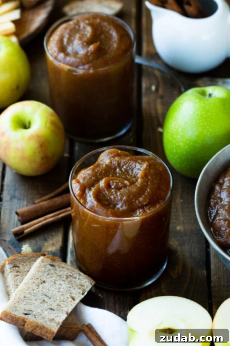 Hands-Off Slow Cooker Apple Butter 2 Two glass jars filled with homemade apple butter, garnished with cinnamon sticks, surrounded by whole fresh apples and peeled apple slices on a rustic surface.