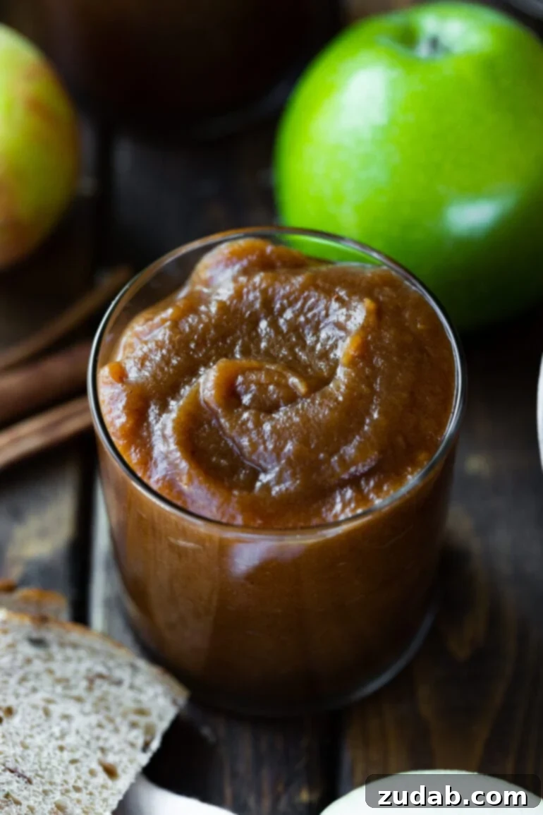 Hands-Off Slow Cooker Apple Butter 4 A glass jar filled with rich, caramel-colored apple butter sits on a wooden surface, with a whole Granny Smith apple in the blurred background.