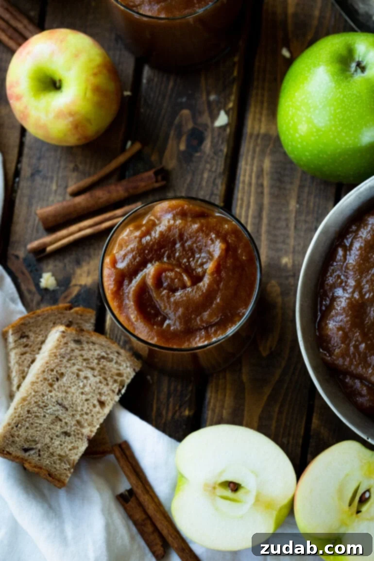 Hands-Off Slow Cooker Apple Butter 6 An overhead shot of a jar of rich apple butter, surrounded by cinnamon sticks, whole apples, halved apples, and a slice of rustic bread, evoking cozy fall vibes.