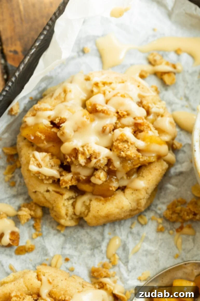 A beautifully baked golden cookie, generously topped with glistening apple pie filling, a crunchy oat crumble, and drizzled with a sweet vanilla glaze. It's the perfect treat for any autumn day.