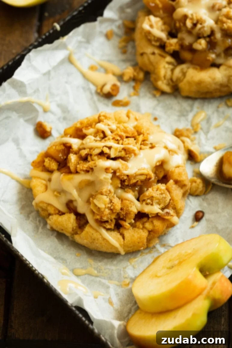 A close-up photo of an apple crisp cookie, showcasing its rich brown sugar cookie base, sweet cinnamon apple filling, and golden, crunchy oat streusel, all finished with a delicate vanilla glaze drizzle.