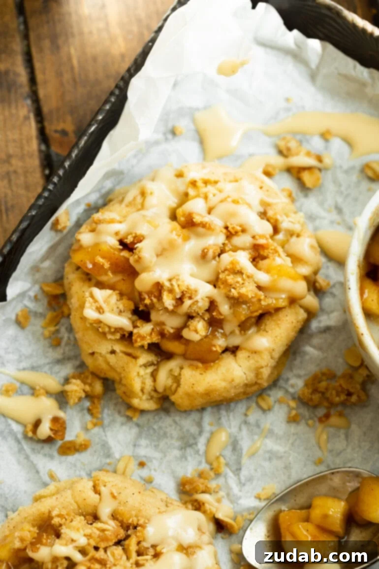A close-up shot of an apple crisp cookie, highlighting its perfectly golden-brown cookie base, the rich and spiced apple filling, the irresistible crunchy oat topping, and a finishing drizzle of vanilla glaze.