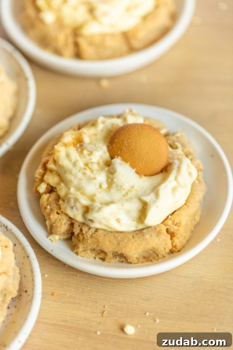 A single large banana cream pie cookie, artfully placed on a small white dessert plate, showcases its golden cookie base and abundant creamy filling.
