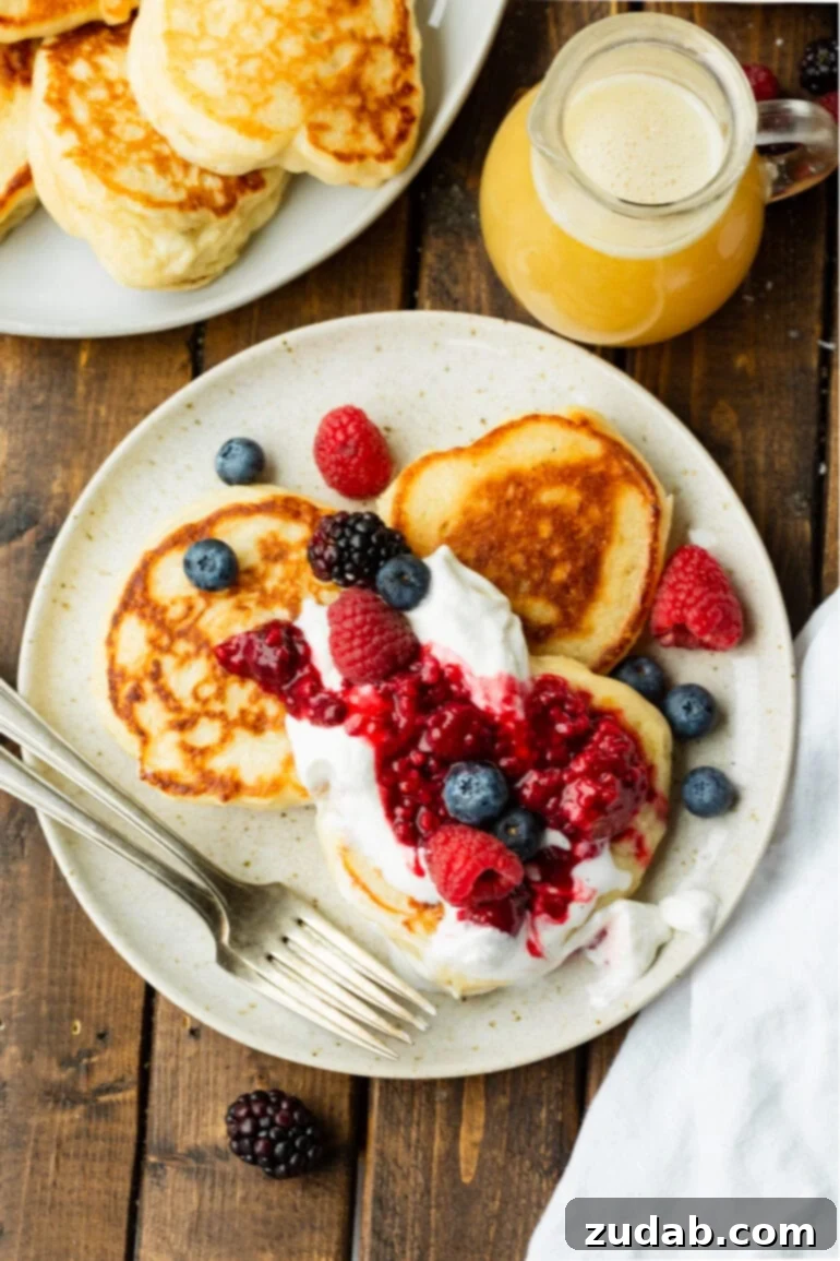 Cloud-Like Fluffy Ukrainian Oladi Pancakes 3 a photo taken over the top of a plate of three golden crispy oladi pancakes topped with mixed fresh berries and whipped cream.