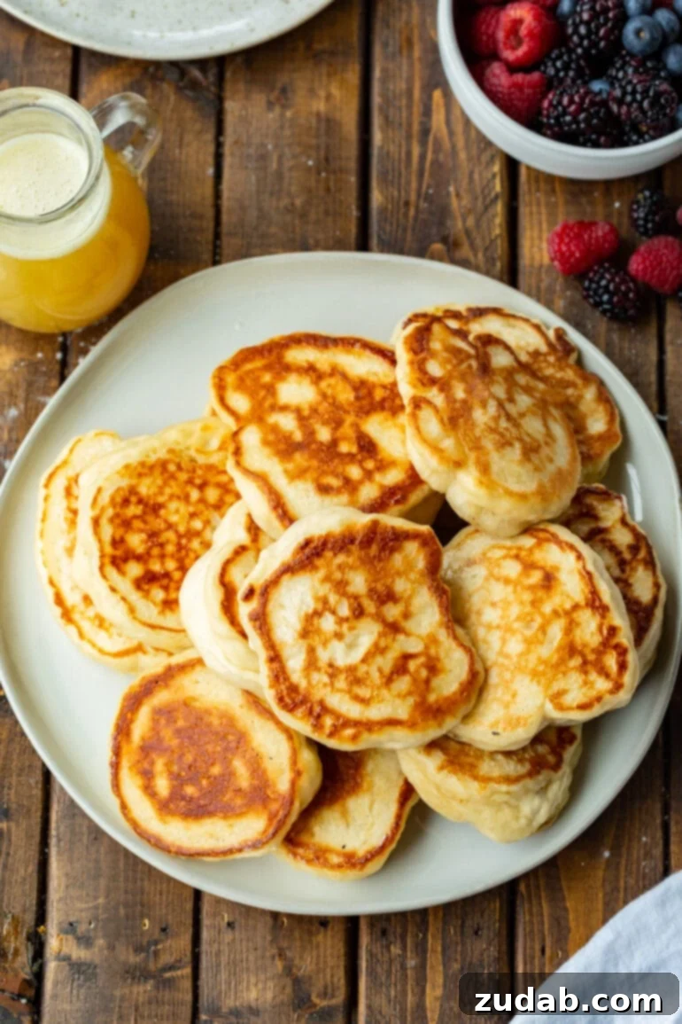 Cloud-Like Fluffy Ukrainian Oladi Pancakes 7 a photo of a large white plate piled up with golden crispy oladi pancakes with a small pitcher of buttermilk syrup sitting next to it.