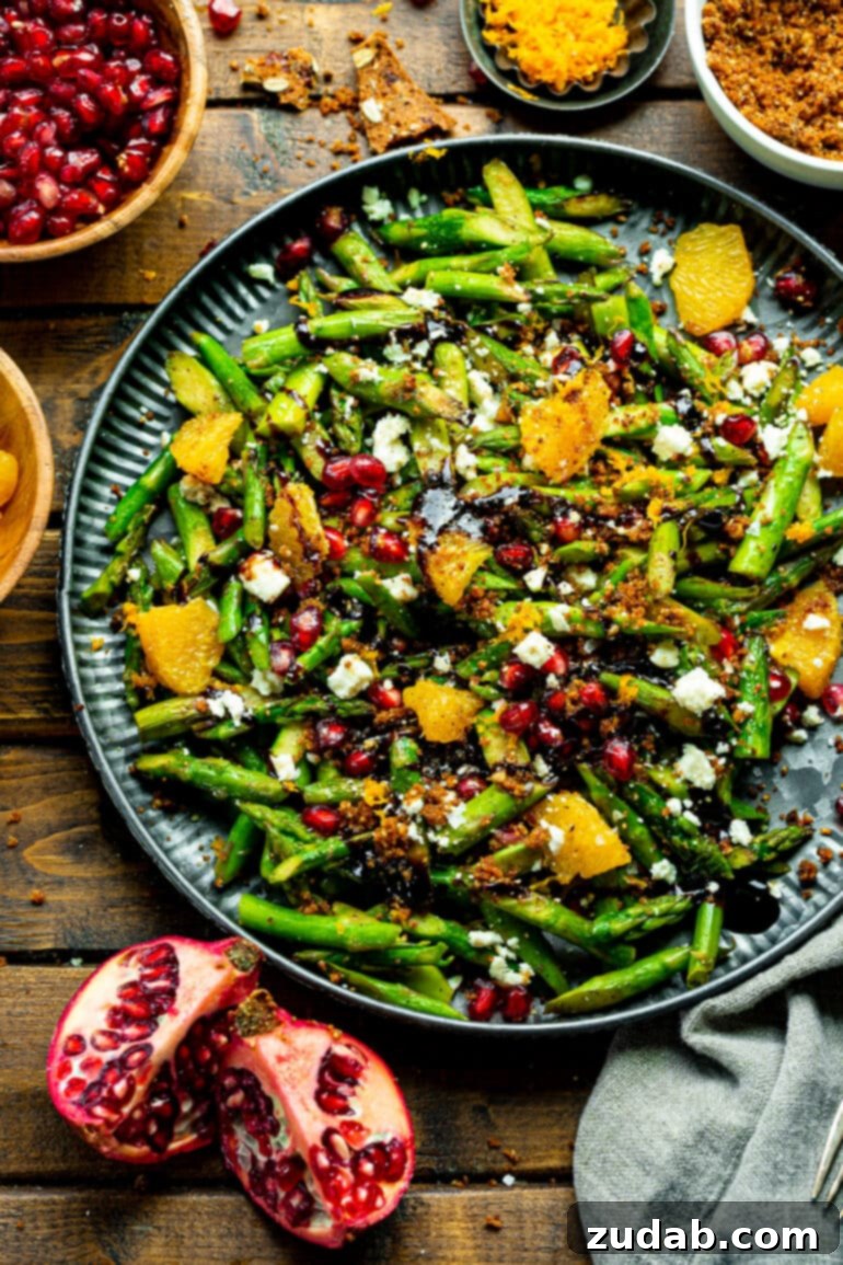 A large serving platter brimming with holiday asparagus salad, generously tossed with bright pomegranate arils, luscious orange segments, crumbled gorgonzola cheese, and a glossy balsamic drizzle.