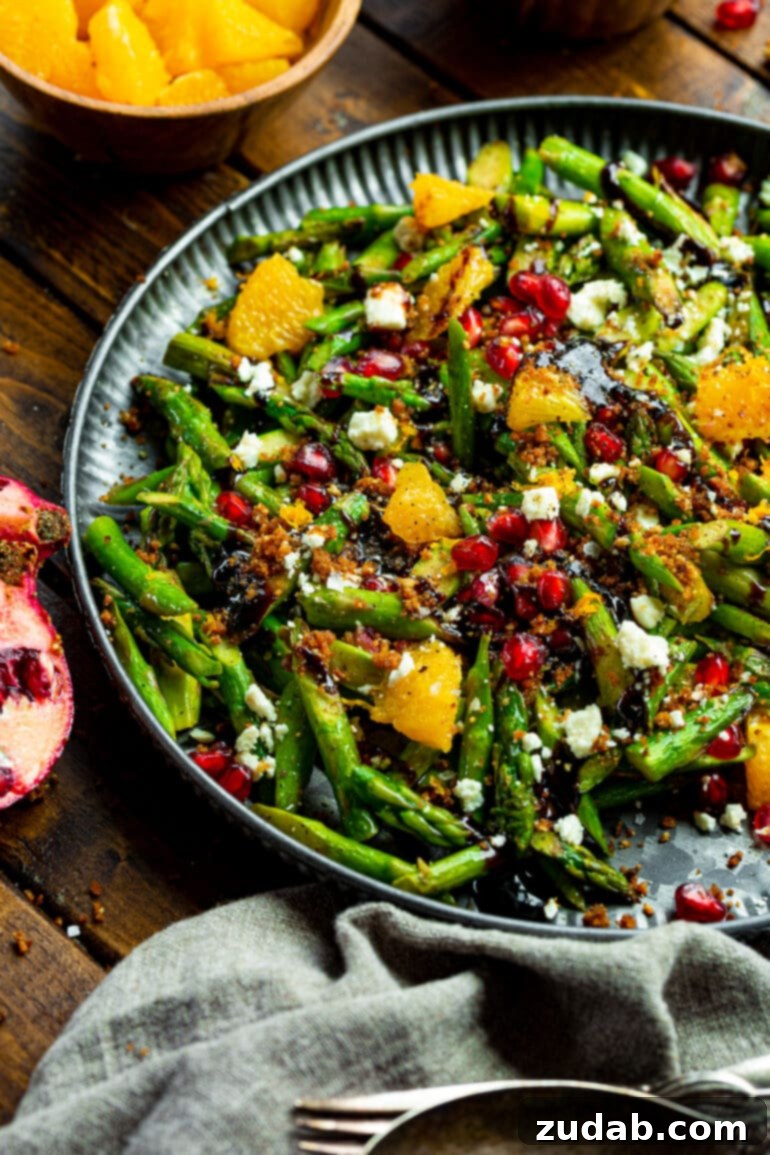 A grand serving tray piled high with a stunning asparagus salad, adorned with ruby-red pomegranates, succulent orange wedges, creamy gorgonzola cheese, perfectly toasted panko breadcrumbs, and a luscious balsamic glaze.