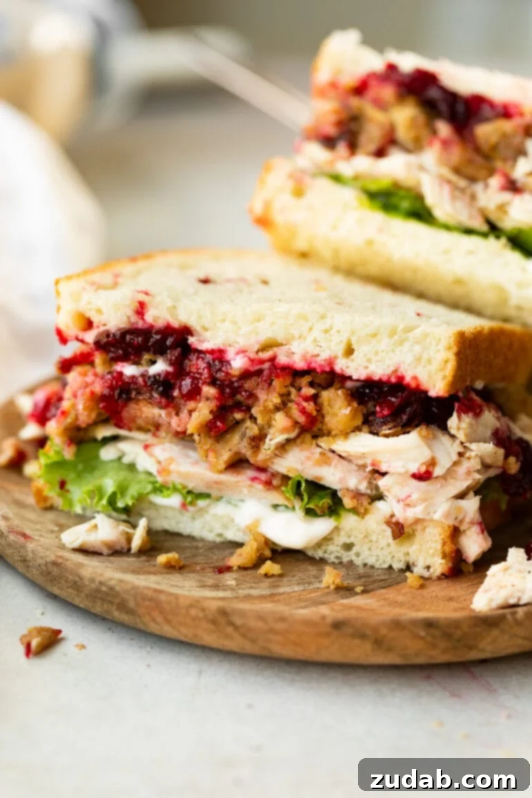 A perfectly assembled half of a Thanksgiving leftover turkey sandwich, known as The Gobbler, showcasing generous layers of roasted turkey, savory stuffing, rich cranberry sauce, crisp lettuce, and a drizzle of gravy, all nestled between slices of white bread. The cross-section reveals the mouthwatering combination of textures and colors, promising a delightful holiday flavor experience.