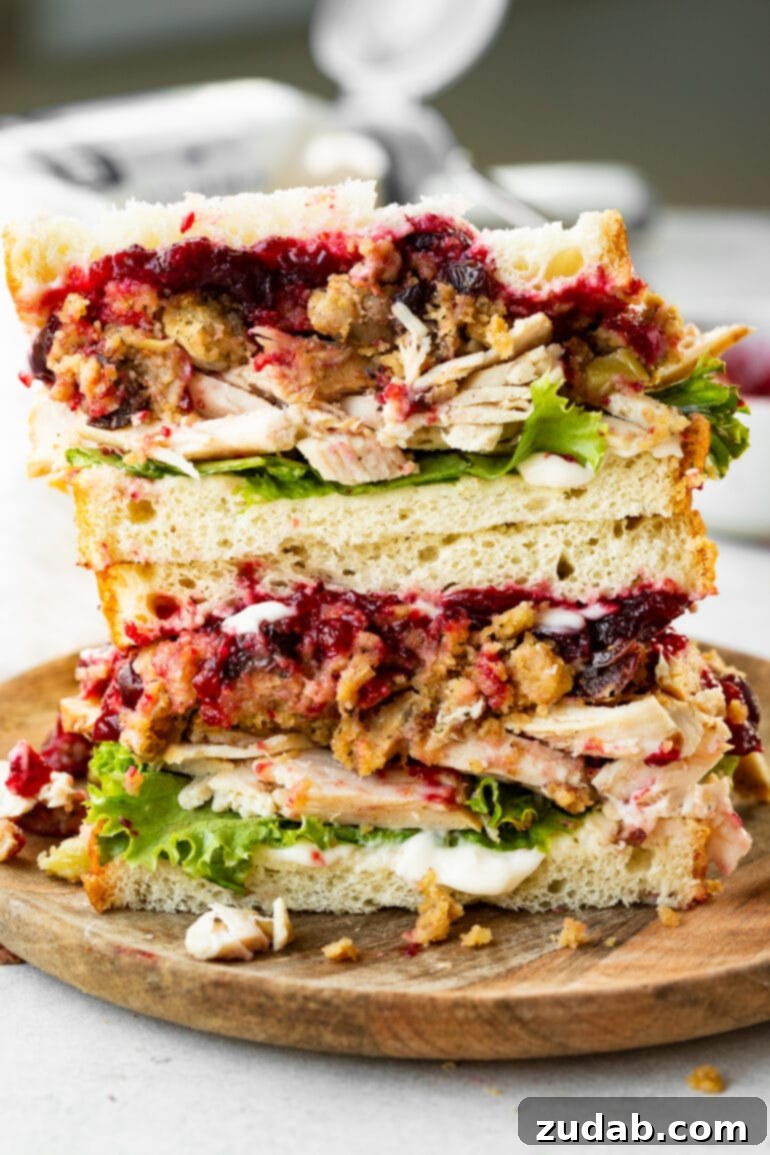 Two halves of The Gobbler, the ultimate Thanksgiving leftover turkey sandwich, are stacked artfully, revealing thick layers of roasted turkey, savory stuffing, and bright cranberry sauce. The golden-brown bread suggests a perfect toast, making it an irresistible post-holiday delight.