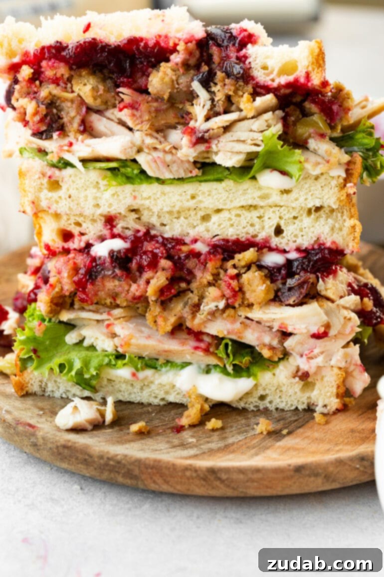 A pair of perfectly sliced Gobbler sandwiches, each half stacked atop the other to beautifully display the cross-section. The layers are clearly visible: tender roasted turkey, crisp shredded lettuce, savory stuffing, rich turkey gravy, and a vibrant layer of cranberry sauce, all expertly contained within soft white bread, promising a full Thanksgiving meal experience in every bite.