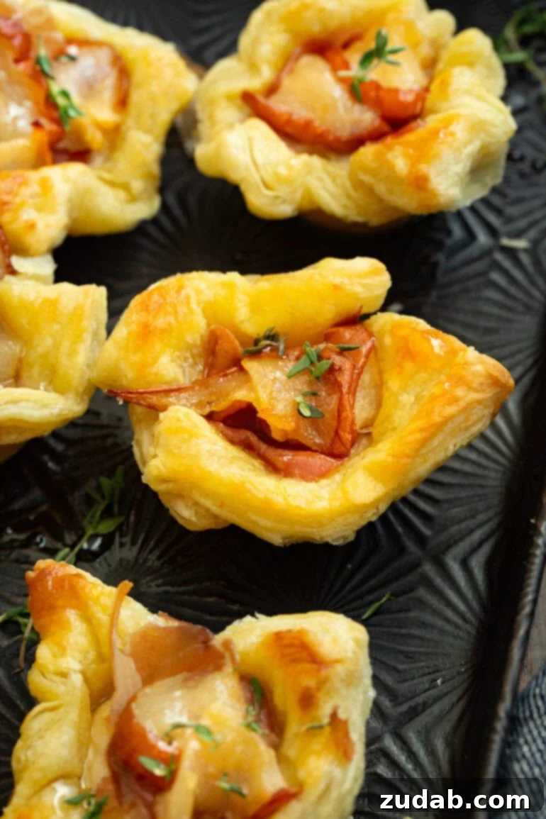 A vibrant photo showcasing several golden-brown puff pastry bites, generously filled with caramelized roasted pear, savory prosciutto, a sprinkle of fresh thyme, and a glistening drizzle of hot honey. Each bite is perfectly sized for an appetizer.