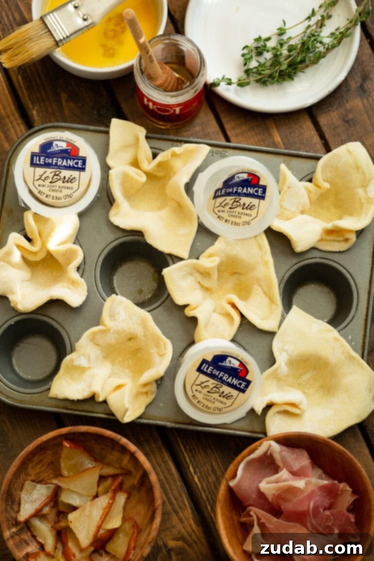 A photo showing a mini muffin tin, with some cups holding unbaked puff pastry squares and others containing small cubes of Brie cheese nestled within the pastry.